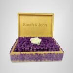 Personalized wooden flower box with "Sarah & John" engraving in Courier New font (2 cm height), filled with vibrant purple real preserved lavender and a single artificial white rose. Ideal for romantic gifts, weddings, anniversaries, or Valentine’s Day celebrations, this handcrafted keepsake box measures 21 cm L x 14 cm W x 7 cm H. The natural wood finish and aromatic lavender scent make it a timeless gift for couples to cherish forever. Crafted with love by Sear Handcrafted Elegance.