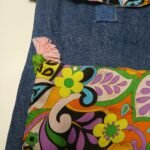 Retro psychedelic denim book bag back view, Sear handcrafted elegance, pink tassel with green pom-pom, vibrant floral fabric, denim top, colorful stitching.