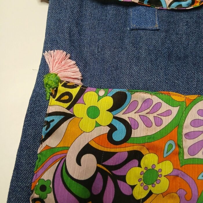 Retro psychedelic denim book bag back view, Sear handcrafted elegance, pink tassel with green pom-pom, vibrant floral fabric, denim top, colorful stitching.