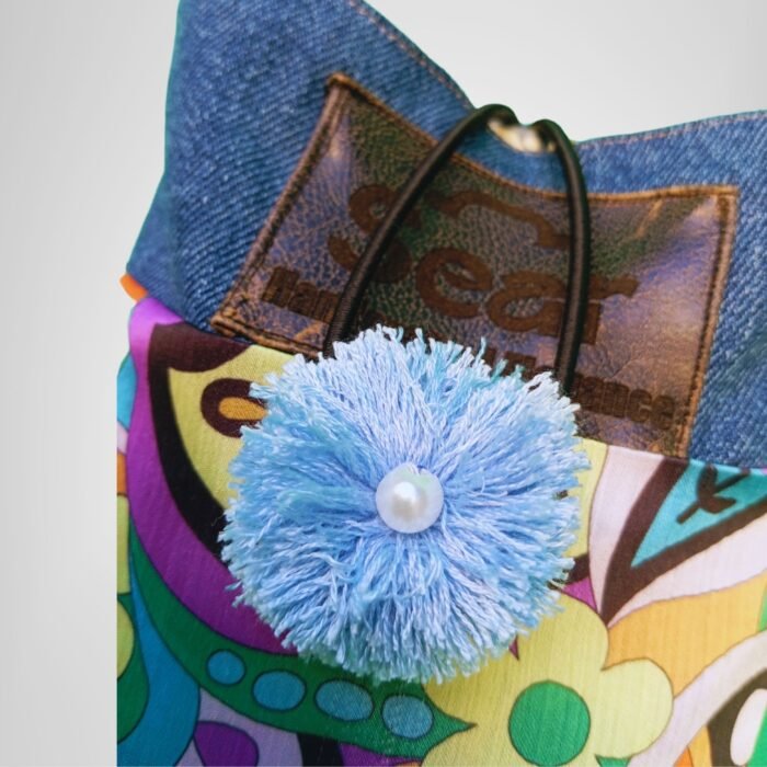 Retro psychedelic denim book bag close-up, Sear handcrafted elegance, blue pom-pom with pearl, vibrant floral fabric, denim top, leather tag.