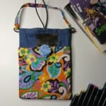 Retro psychedelic denim book bag with blue pom-pom, Sear handcrafted elegance, vibrant floral fabric, denim top, colorful strap, books, markers nearby.