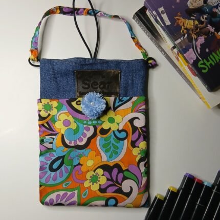 Retro psychedelic denim book bag with blue pom-pom, Sear handcrafted elegance, vibrant floral fabric, denim top, colorful strap, books, markers nearby.