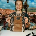 Sema Arslan holds a leopard print book organizer bag with a brown pocket by Sear Handcrafted Elegance, showcasing its stylish design. The background features a vibrant outdoor setting with a scenic view of a hilly town under a partly cloudy sky, framed by decorative lanterns, a tiled floor, and a small fountain. Books, glasses, and a pen are placed on the white table in the foreground.