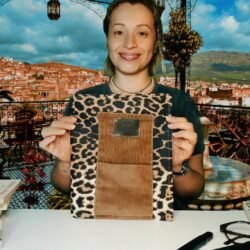 Sema Arslan holds a leopard print book organizer bag with a brown pocket by Sear Handcrafted Elegance, showcasing its stylish design. The background features a vibrant outdoor setting with a scenic view of a hilly town under a partly cloudy sky, framed by decorative lanterns, a tiled floor, and a small fountain. Books, glasses, and a pen are placed on the white table in the foreground.