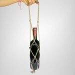 A Sear Handcrafted Elegance macrame wine bottle bag in brown, handmade from 100% recycled cotton, holding a wine bottle. The bag features an intricate knot design with brown decorative beads and a tassel at the bottom, held by a hand against a plain background.