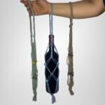 A Sear Handcrafted Elegance macrame wine bottle bag, handmade from 100% recycled cotton, displayed in three colors: green, blue, and beige. The blue bag holds a wine bottle, showcasing the intricate knot design with decorative beads. A hand holds the bags against a plain background.
