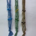 Three Sear Handcrafted Elegance macrame wine bottle bags, handmade from 100% recycled cotton, in blue, green, and beige. Each bag features an intricate knot design with decorative beads in blue, green, and brown, hanging against a plain background.