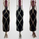 A Sear Handcrafted Elegance sim macrame wine bottle bag, handmade with shimmering threads, displayed in three colors: gold, silver, and red. Each bag holds a wine bottle, showcasing intricate knot designs with a sparkling finish and tassels, against a plain background.