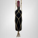 A Sear Handcrafted Elegance sim macrame wine bottle bag in gold, handmade with shimmering threads, holding a wine bottle. The bag features an intricate knot design with a sparkling finish and a tassel at the bottom, displayed against a plain background.