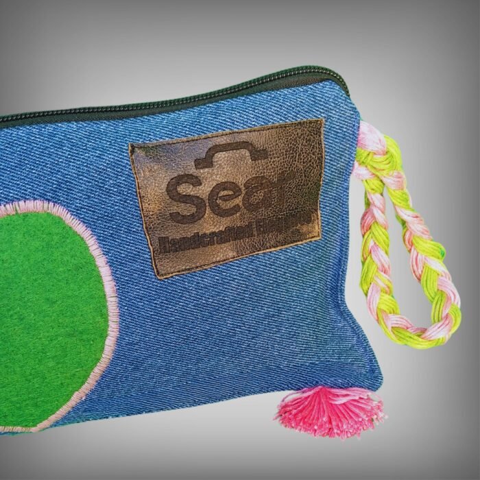 Close-up of a blue denim makeup bag by Sear Handcrafted Elegance, highlighting the green heart design with pink stitching and the pink-green braided strap detail. A trendy and functional cosmetic bag for beauty essentials.