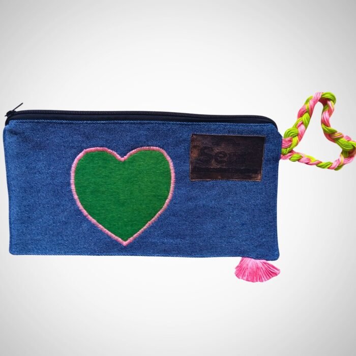 A stylish blue denim makeup bag by Sear Handcrafted Elegance, featuring a green heart design with pink stitching, a pink and green braided strap, and a pink tassel detail. This zippered cosmetic pouch is perfect for organizing beauty essentials.