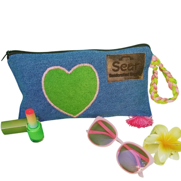 A stylish blue denim makeup bag by Sear Handcrafted Elegance, featuring a green heart design with pink stitching, a pink and green braided strap, and a pink tassel detail. This zippered cosmetic pouch is perfect for organizing beauty essentials.