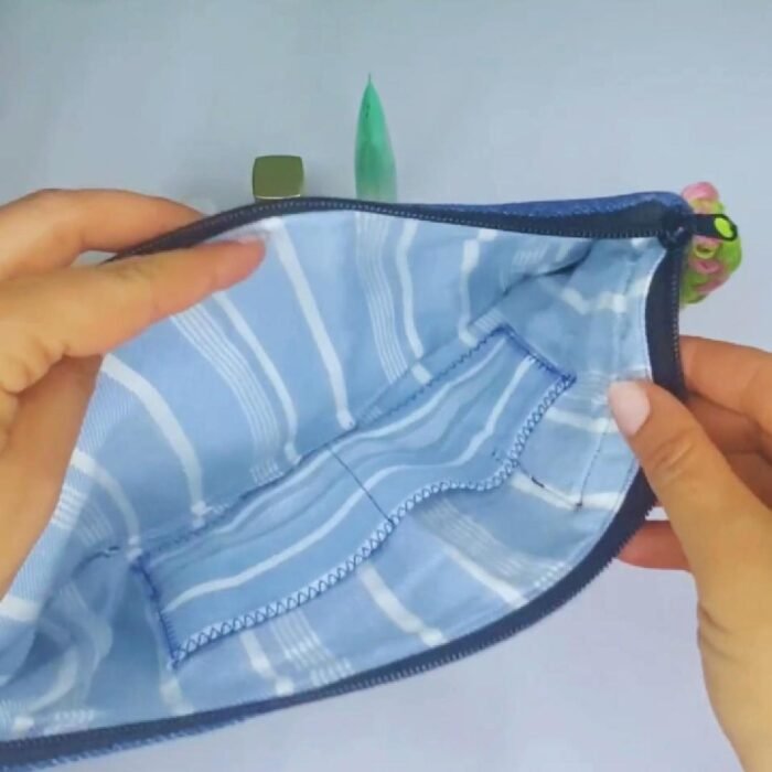 A stylish blue denim makeup bag by Sear Handcrafted Elegance, featuring a green heart design with pink stitching, a pink and green braided strap, and a pink tassel detail. This zippered cosmetic pouch is perfect for organizing beauty essentials.