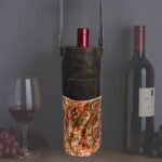 Handmade leather paisley wine bag with leather strap by Sear Handcrafted Elegance, holding a wine bottle, displayed with a wine glass and grapes on a dark background.
