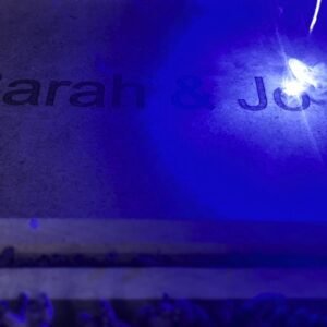 sear-engraving-personalized-text-with-laser-cnc Close-up of a laser CNC machine engraving the personalized text 'Sarah & John' onto a flat surface, illuminated by a bright blue laser light, with smoke and sparks visible during the process.