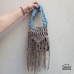 A stylish handmade macrame handbag by Sear Handcrafted Elegance, featuring intricate beige macrame weaving, a blue handle, and a leopard print linen base with long fringes, held against a textured white wall.