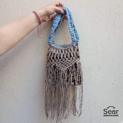 A stylish handmade macrame handbag by Sear Handcrafted Elegance, featuring intricate beige macrame weaving, a blue handle, and a leopard print linen base with long fringes, held against a textured white wall.
