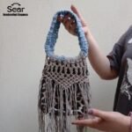 A stylish handmade macrame handbag by Sear Handcrafted Elegance, featuring intricate beige macrame weaving, a blue handle, and a leopard print linen base with long fringes, held against a textured white wall. Video Cover.