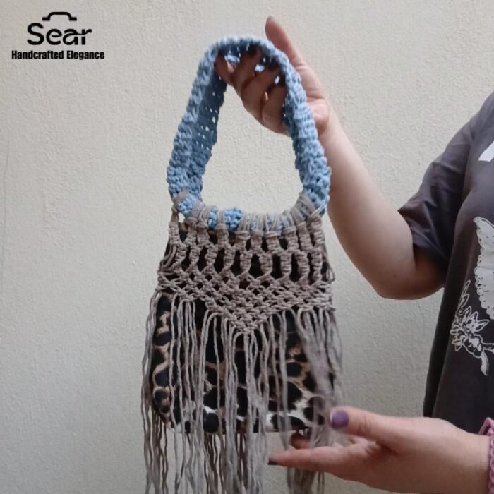 A stylish handmade macrame handbag by Sear Handcrafted Elegance, featuring intricate beige macrame weaving, a blue handle, and a leopard print linen base with long fringes, held against a textured white wall. Video Cover.