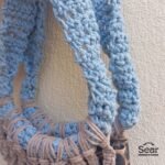 A side view of a handmade macrame handbag by Sear Handcrafted Elegance, featuring a blue macrame handle and beige weaving details with a leopard print linen base, against a textured white wall.