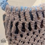 A close-up view of a handmade macrame handbag by Sear Handcrafted Elegance, showcasing detailed beige macrame weaving, a blue handle, and a leopard print linen base against a textured white wall.