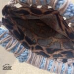 An inside view of a handmade macrame handbag by Sear Handcrafted Elegance, featuring a leopard print linen lining, beige macrame weaving, and a blue handle, against a textured white wall.