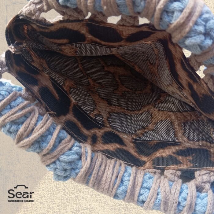 An inside view of a handmade macrame handbag by Sear Handcrafted Elegance, featuring a leopard print linen lining, beige macrame weaving, and a blue handle, against a textured white wall.
