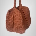 An angled view of the Sear Handcrafted Elegance terracotta crochet tote bag, displaying its warm, earthy brown shade and expertly crafted crochet texture. The bag features a practical handle and a playful tassel detail, adding a bohemian touch to its elegant design. Perfect for casual outings or as a statement piece, this handmade tote blends artisan craftsmanship with timeless style. The soft lighting and neutral gray background highlight the bag’s intricate stitching and rustic charm.