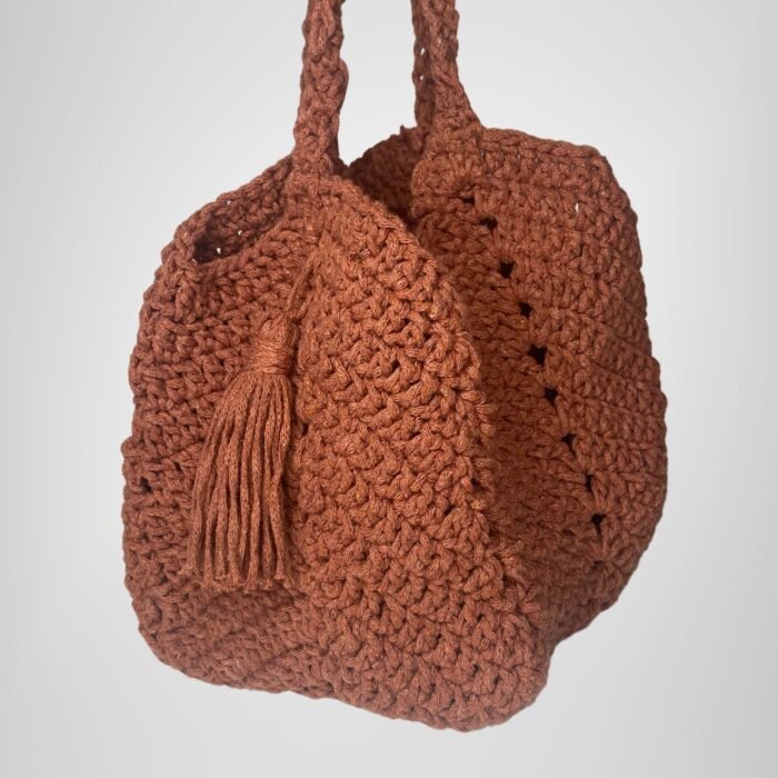 An angled view of the Sear Handcrafted Elegance terracotta crochet tote bag, displaying its warm, earthy brown shade and expertly crafted crochet texture. The bag features a practical handle and a playful tassel detail, adding a bohemian touch to its elegant design. Perfect for casual outings or as a statement piece, this handmade tote blends artisan craftsmanship with timeless style. The soft lighting and neutral gray background highlight the bag’s intricate stitching and rustic charm.