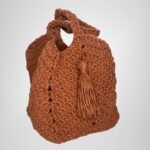 An angled view of the Sear Handcrafted Elegance terracotta crochet tote bag, displaying its warm, earthy brown shade and expertly crafted crochet texture. The bag features a practical handle and a playful tassel detail, adding a bohemian touch to its elegant design. Perfect for casual outings or as a statement piece, this handmade tote blends artisan craftsmanship with timeless style. The soft lighting and neutral gray background highlight the bag’s intricate stitching and rustic charm.