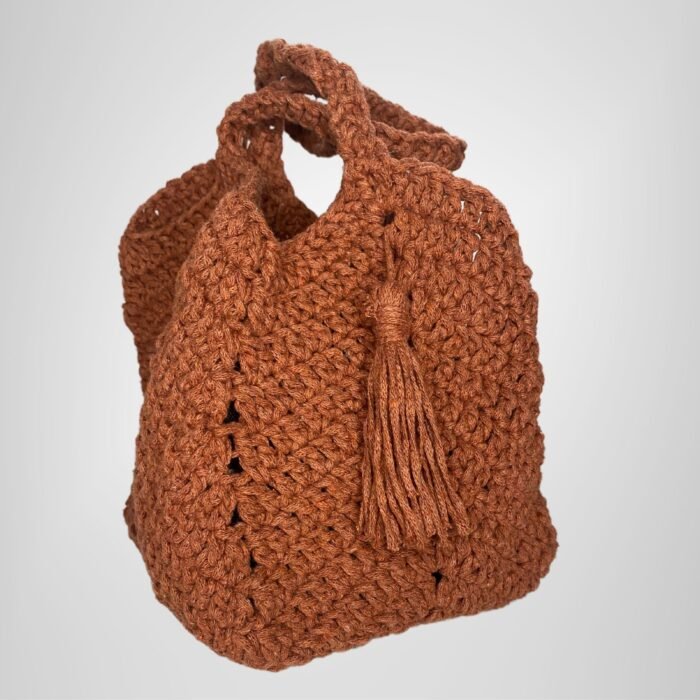 An angled view of the Sear Handcrafted Elegance terracotta crochet tote bag, displaying its warm, earthy brown shade and expertly crafted crochet texture. The bag features a practical handle and a playful tassel detail, adding a bohemian touch to its elegant design. Perfect for casual outings or as a statement piece, this handmade tote blends artisan craftsmanship with timeless style. The soft lighting and neutral gray background highlight the bag’s intricate stitching and rustic charm.
