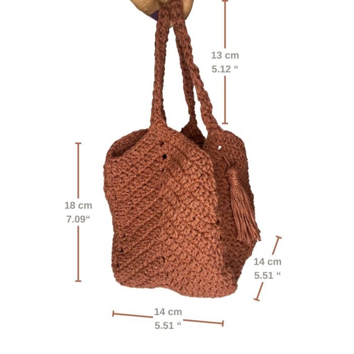 A detailed dimensions view of the Sear Handcrafted Elegance terracotta crochet tote bag, showcasing its warm brown hue and intricate crochet design. The bag measures 18 cm (7.09") in height, 14 cm (5.51") in width, and 13 cm (5.12") in handle drop, as indicated by labeled measurements. Featuring a tassel accent, this handmade tote combines rustic style with practical sizing, perfect for everyday use. The bag is held by a hand against a white background, highlighting its textured craftsmanship.
