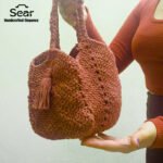 A person holding the Sear Handcrafted Elegance terracotta crochet tote bag, showcasing its rich, earthy brown tone and intricate crochet texture. The bag features a sturdy handle and a decorative tassel, adding a bohemian flair to its elegant design. Perfect for everyday use or as a stylish accessory, this handmade tote is presented against a soft, light background with the "Sear Handcrafted Elegance" logo in the top left corner, emphasizing its artisan craftsmanship.