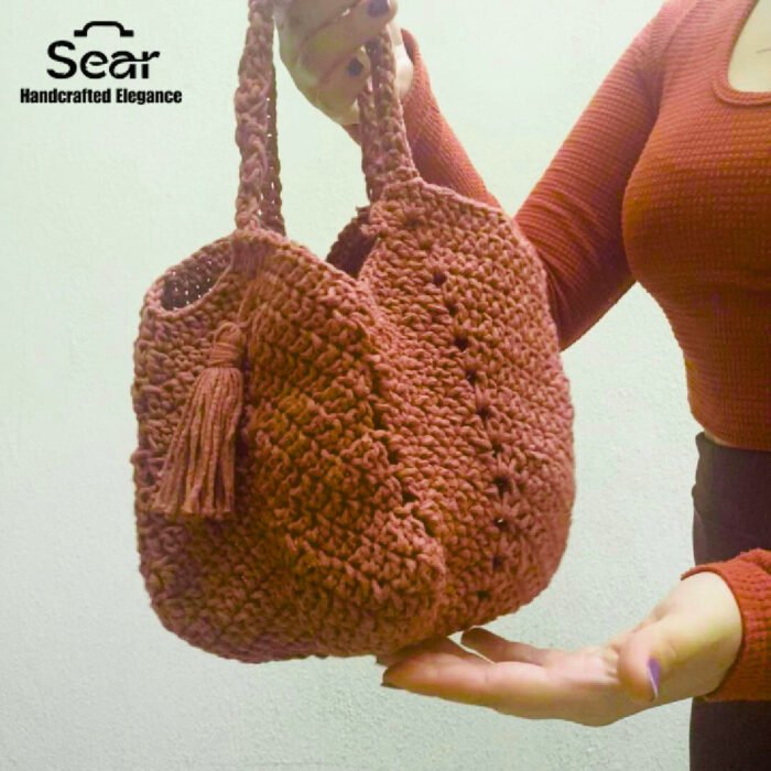 A person holding the Sear Handcrafted Elegance terracotta crochet tote bag, showcasing its rich, earthy brown tone and intricate crochet texture. The bag features a sturdy handle and a decorative tassel, adding a bohemian flair to its elegant design. Perfect for everyday use or as a stylish accessory, this handmade tote is presented against a soft, light background with the "Sear Handcrafted Elegance" logo in the top left corner, emphasizing its artisan craftsmanship.