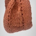 A side view of the Sear Handcrafted Elegance terracotta crochet tote bag, highlighting its warm, rustic brown hue and detailed crochet pattern. The bag features a charming tassel detail on the front, adding a touch of bohemian flair. Crafted with care, this handmade tote combines style and practicality, making it a versatile accessory for daily use or special occasions. The textured stitching and soft, earthy tones are beautifully showcased against a simple gray background.
