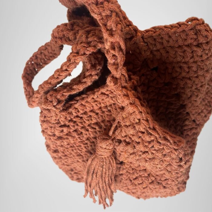 A top-angled view of the Sear Handcrafted Elegance terracotta crochet tote bag, revealing its warm brown tones and detailed crochet craftsmanship. The bag features a sturdy handle and a delicate tassel accent, enhancing its boho-inspired aesthetic. Ideal for carrying essentials with style, this handmade tote combines rustic charm with sophisticated design. The soft gray background emphasizes the bag’s textured stitching and earthy elegance, making it a perfect accessory for any occasion.