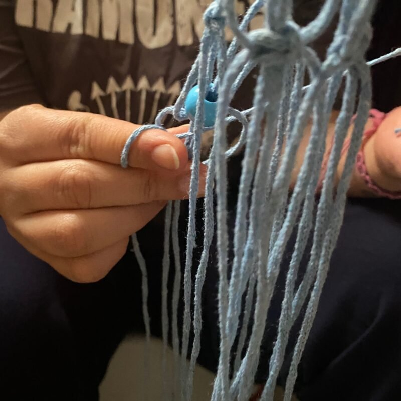 Sema, co-founder of Sear Handcrafted Elegance, crafting a macramé wine or bottle bag with light blue yarn and a turquoise bead, wearing a dark 'RAMONES' shirt